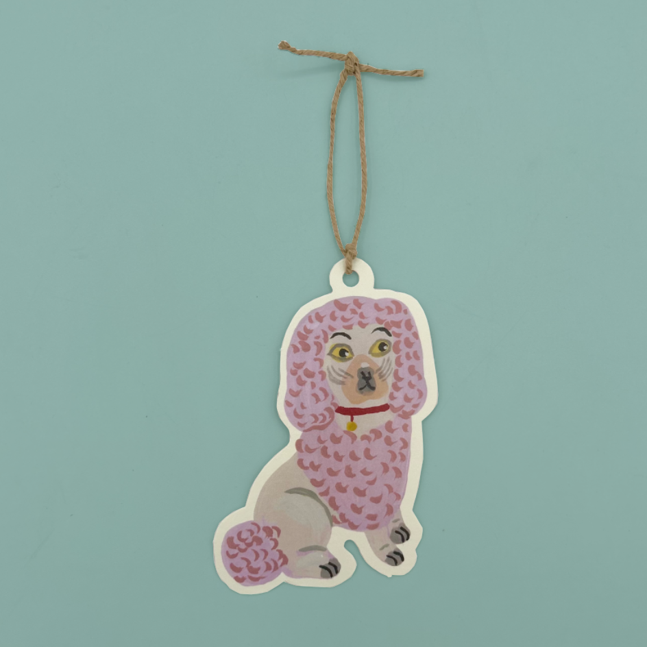 A die-cut hanging gift tag featuring a pink poodle with yellow eyes, and a red collar, suspended by a natural jute string against a solid light teal background.