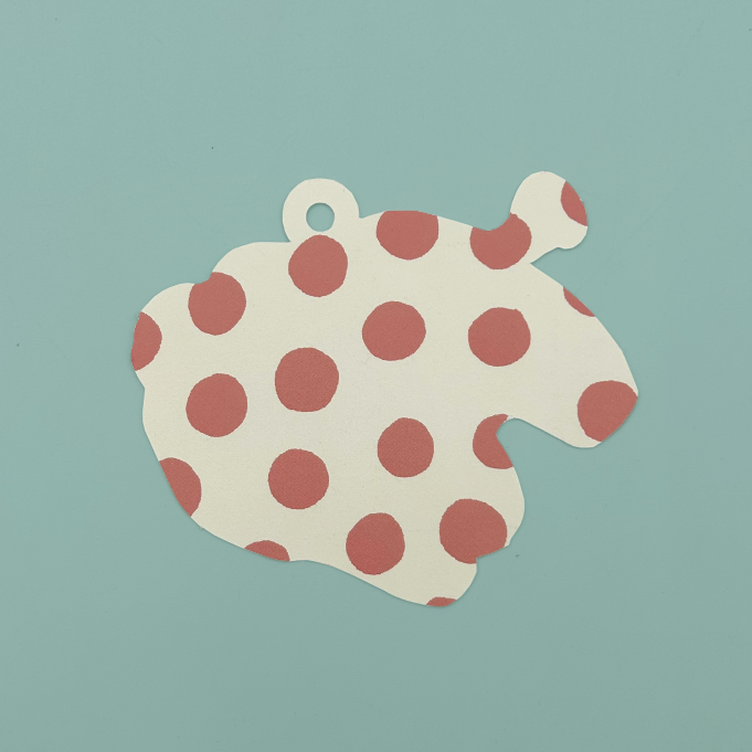 The back of a die cut gift tag with a small hanging loop at the top, covered in evenly spaced dusty rose polka dots, shown on a solid muted teal background.