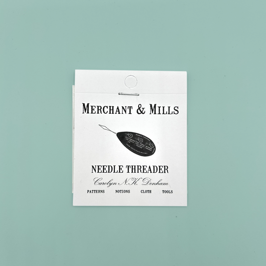 Merchant & Mills needle threader white cardboard packaging which is held together with a staple. On a blue background. 