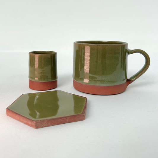Three-piece ceramic tableware set including a hexagonal coaster, an egg cup, and a mug, all glazed in olive green with a terracotta-colored unglazed base.