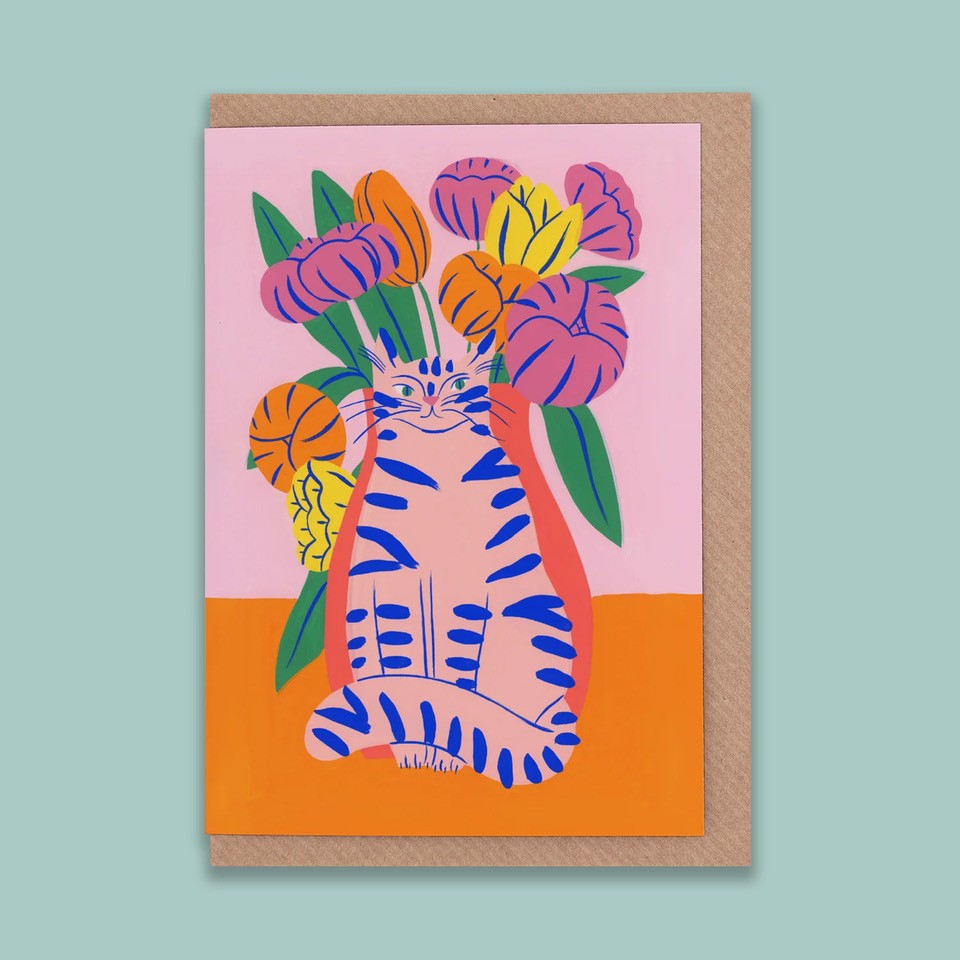 A portrait orientation greetings card with a digital illustration of a pink cat in front of pink, yellow, and orange flowers on a pink and orange background. The card is on a brown envelope on a plain backdrop. 