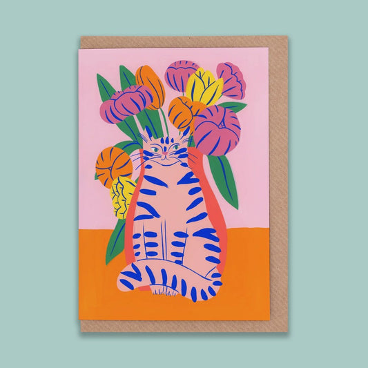 A portrait orientation greetings card with a digital illustration of a pink cat in front of pink, yellow, and orange flowers on a pink and orange background. The card is on a brown envelope on a plain backdrop. 