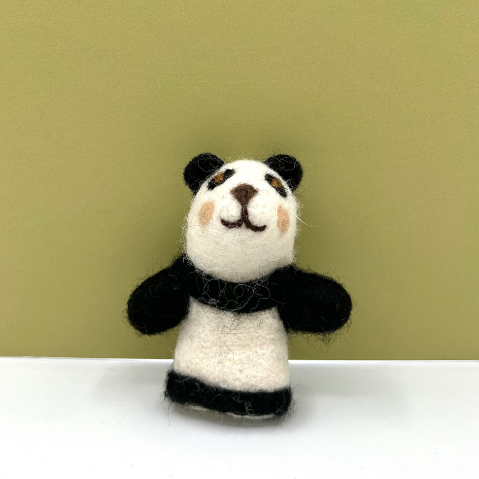 A small felted wool panda finger puppet with black and white fur, embroidered facial features, and a black scarf, standing on a white surface against a muted olive-green background.