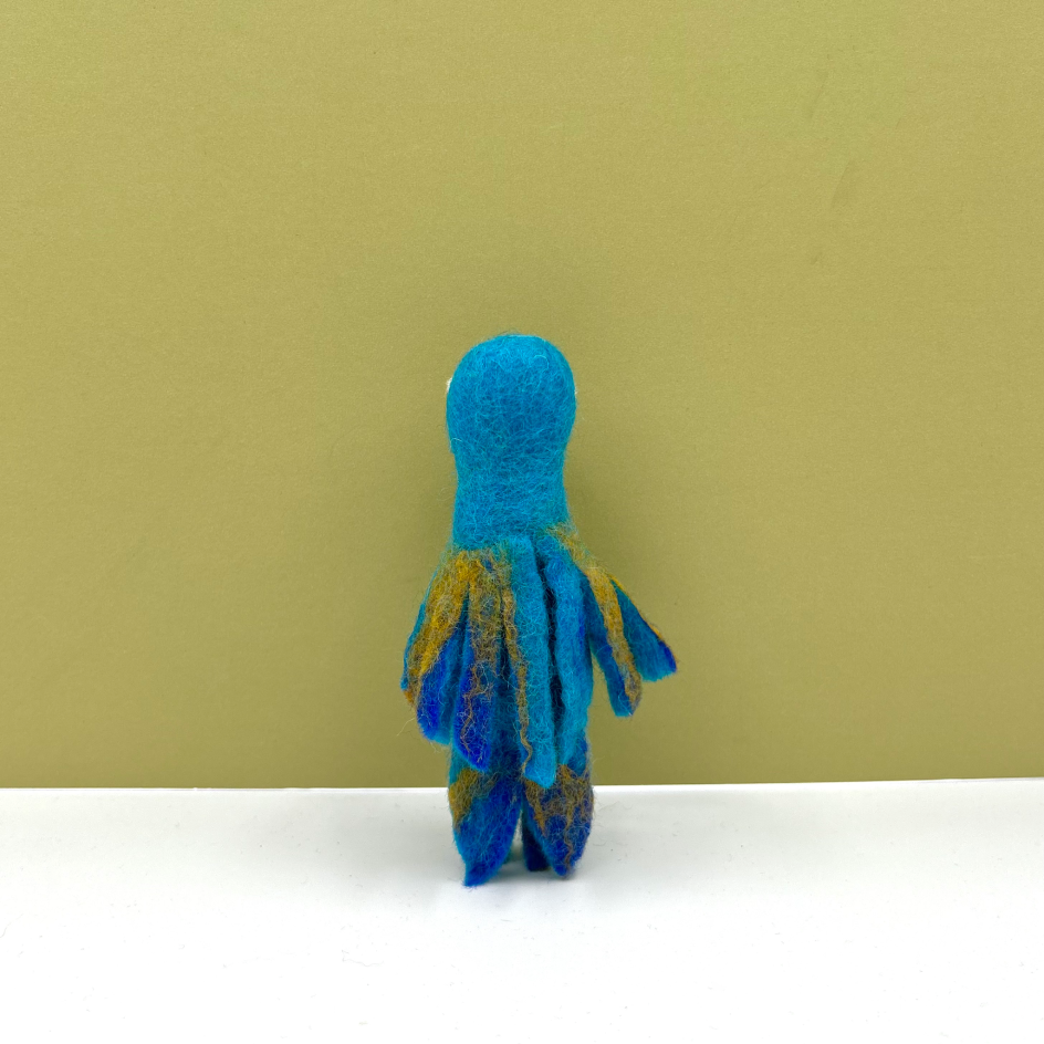 A small felt parrot puppet shown from the back, featuring a bright blue body and darker blue and gold striped feathers, set against a solid olive-green background on a white surface.