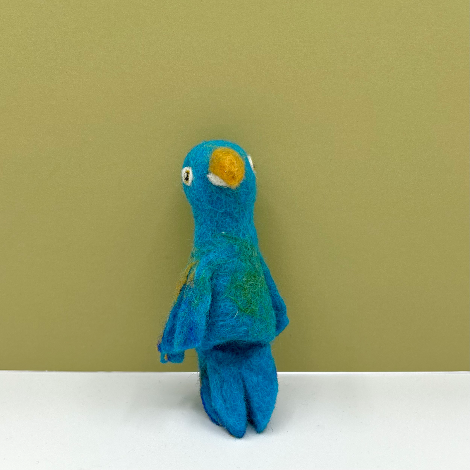 A small felted wool parrot puppet in vibrant blue with a yellow beak and subtle green feather accents, standing on a white surface against a solid olive green background.