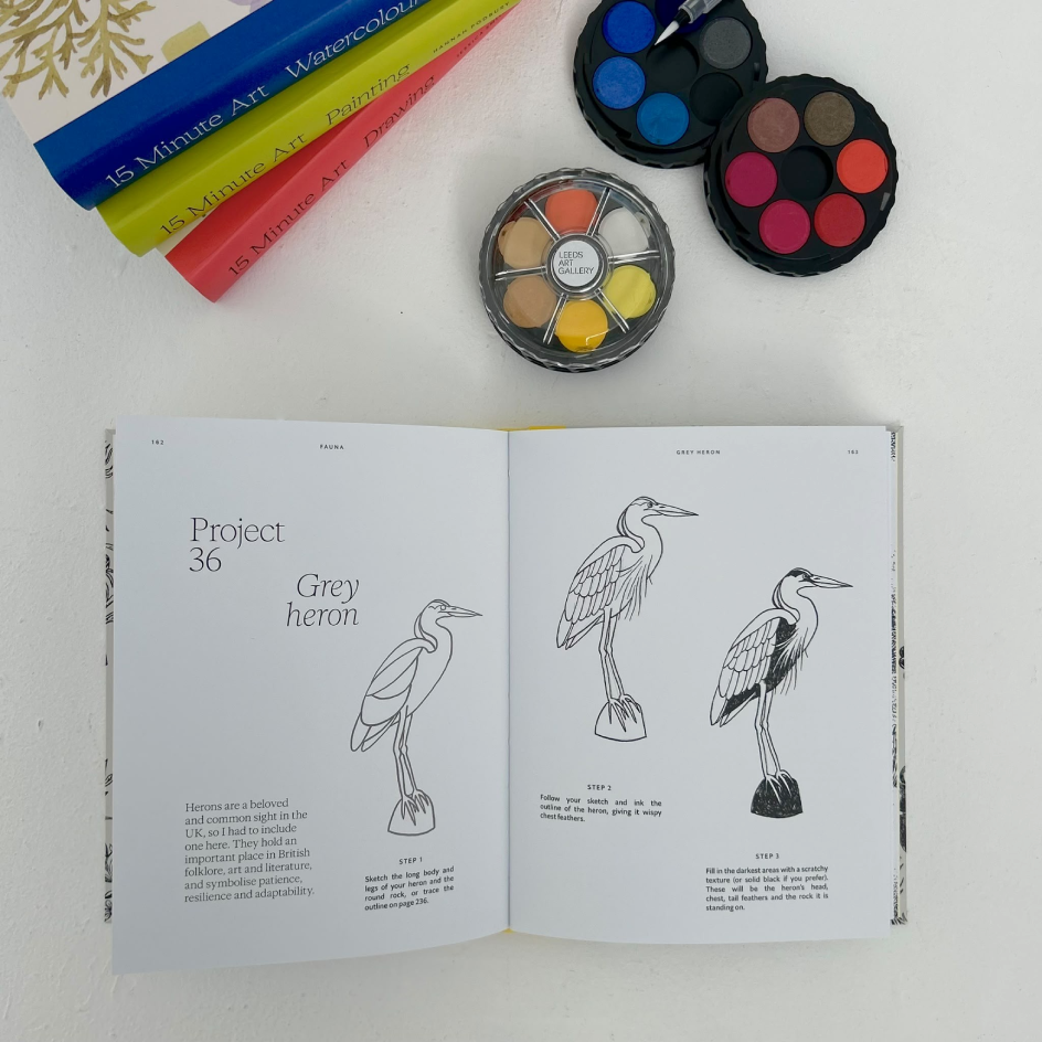 An open watercolor painting book showing step-by-step instructions for painting a grey heron, displayed on a white surface alongside watercolour  paint palettes.