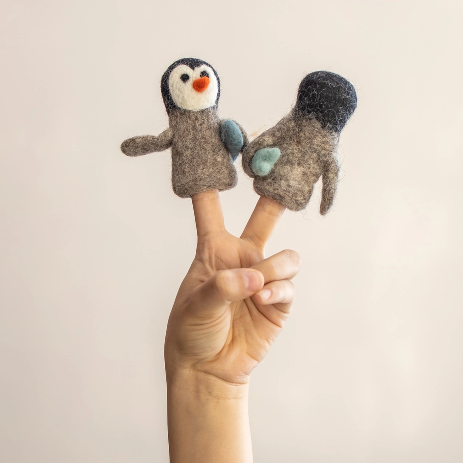 A hand holding two felt penguin finger puppets against a plain light beige background. 