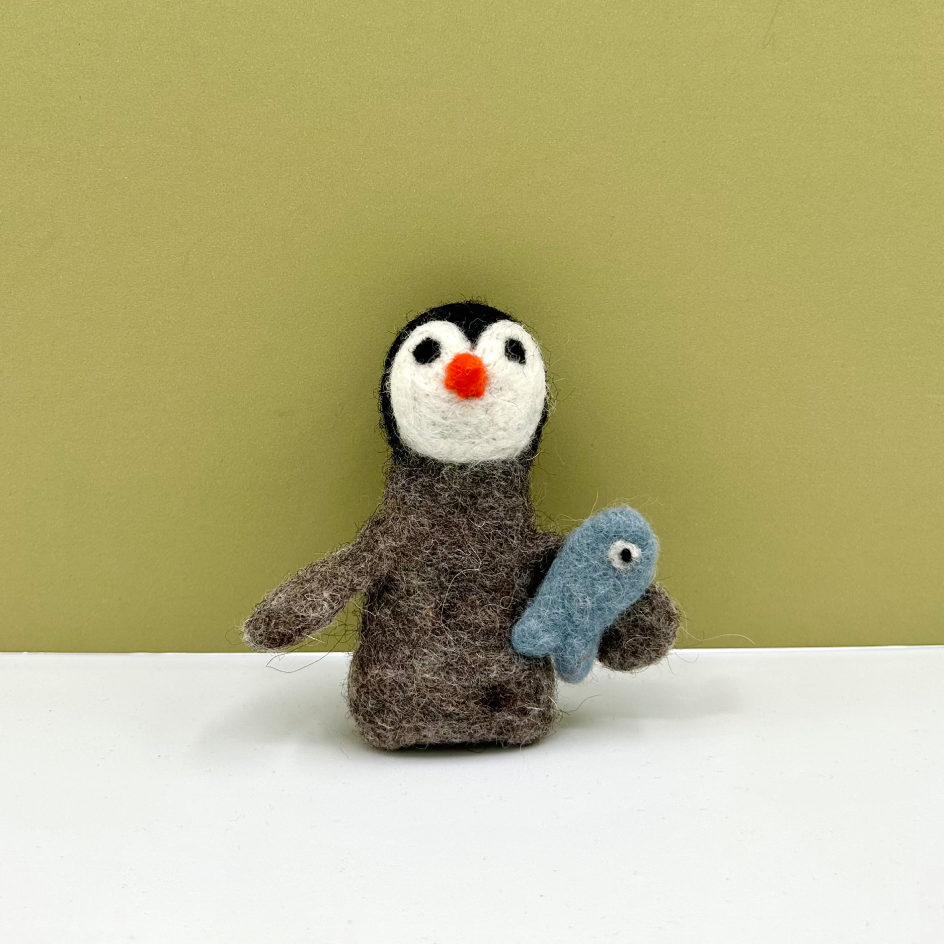 A handmade felted wool penguin toy holding a small blue fish, standing on a white surface against a solid olive green background.