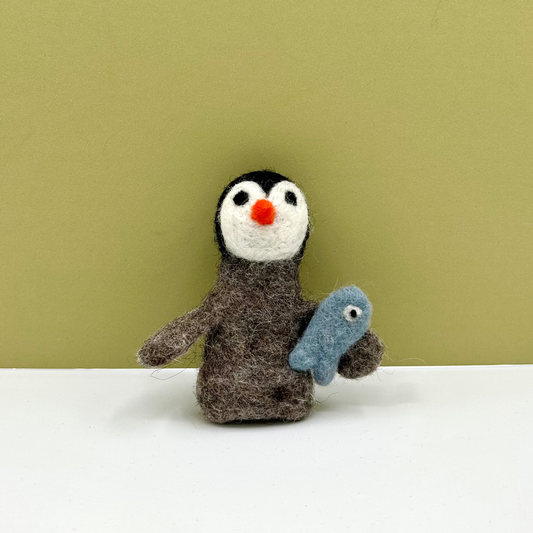 A handmade felted wool penguin toy holding a small blue fish, standing on a white surface against a solid olive green background.