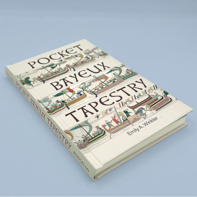 A pocket-size book titled 'Bayeux Tapestry' by Emily A. Winkler, featuring a detailed reproduction of the famous medieval tapestry on the cover, photographed on a light blue background.