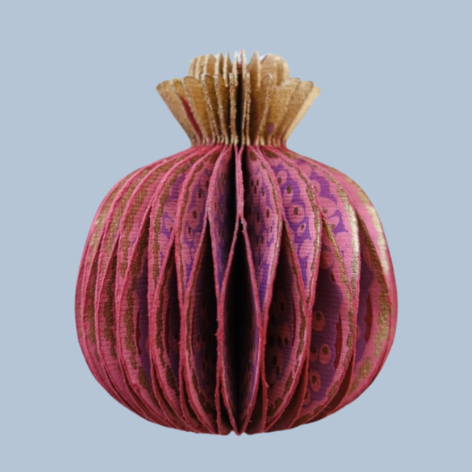 A decorative pomegranate ornament made from layered purple and pink patterned paper with a gold-toned top, shown against a solid light blue background.
