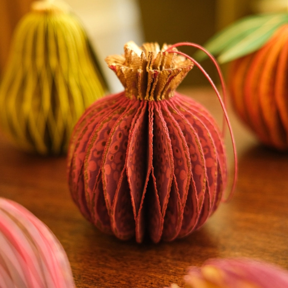A close-up of a decorative paper ornament shaped like a pomegranate, made from layered red patterned paper with a gold-toned textured top and a thin red hanging string, set on a wooden surface with other colorful ornaments blurred in the background.