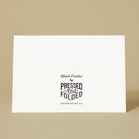 The back of a white rectangular card  standing upright against a warm beige background. The center features black text reading 'Hand Printed by PRESSED And FOLDED' with the website 'pressedandfolded.