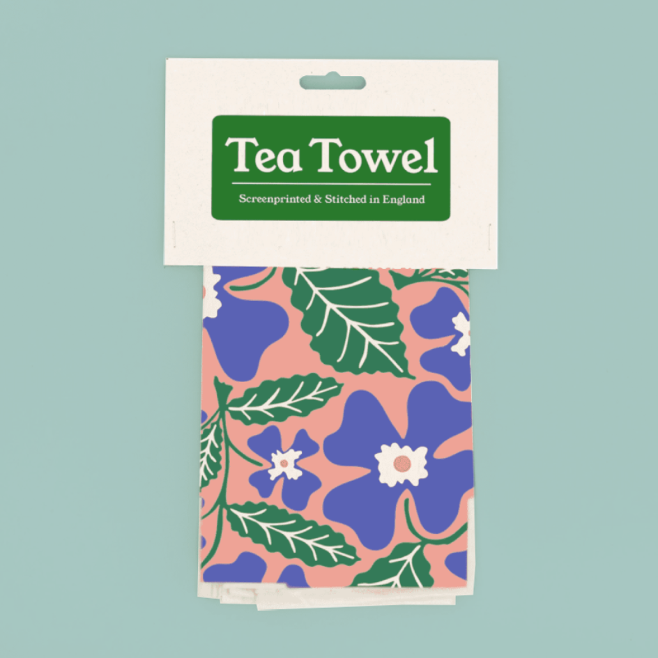 A packaged tea towel with a retro floral pattern in pink, purple, green, and white, hanging from a white paper header card with a green label that reads 'Tea Towel'.