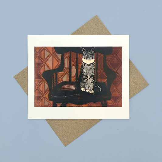 Greetings card with a linocut print of a cat sat on a chair in front of orange wallpaper. The card is on a brown envelope on a blue background.