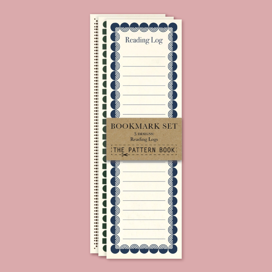 A set of three spiral-bound reading logs from The Pattern Book, stacked vertically on a dusty pink background. The logs are cream-colored with navy blue decorative borders and a brown kraft paper band wrapped around the middle that reads 'BOOKMARK SET 3 READING Reading Logs THE PATTERN BOOK'.