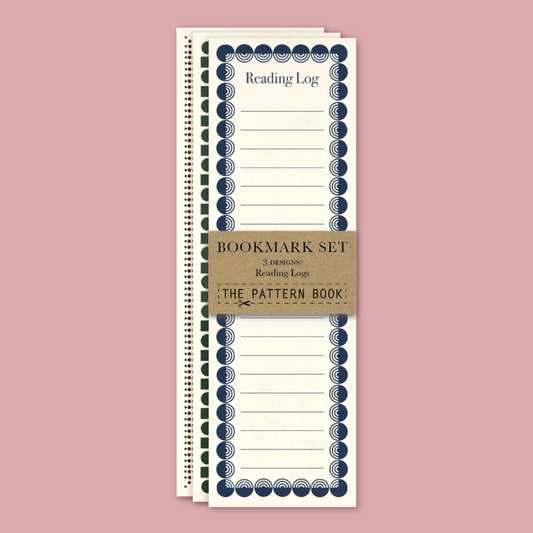 A set of three spiral-bound reading logs from The Pattern Book, stacked vertically on a dusty pink background. The logs are cream-colored with navy blue decorative borders and a brown kraft paper band wrapped around the middle that reads 'BOOKMARK SET 3 READING Reading Logs THE PATTERN BOOK'.