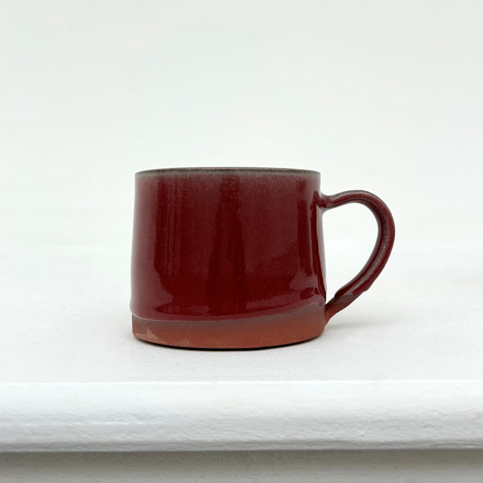 A ceramic mug with a terracotta coloured base and a burgundy red glossy glaze. The mug is a on a white ledge with a white wall behind.