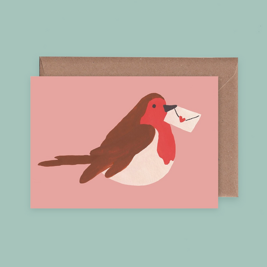 A pink greetings card with a pinted illustration of a robin holding a letter in its beak. The card is against a brown envelope on a pale teal background. 