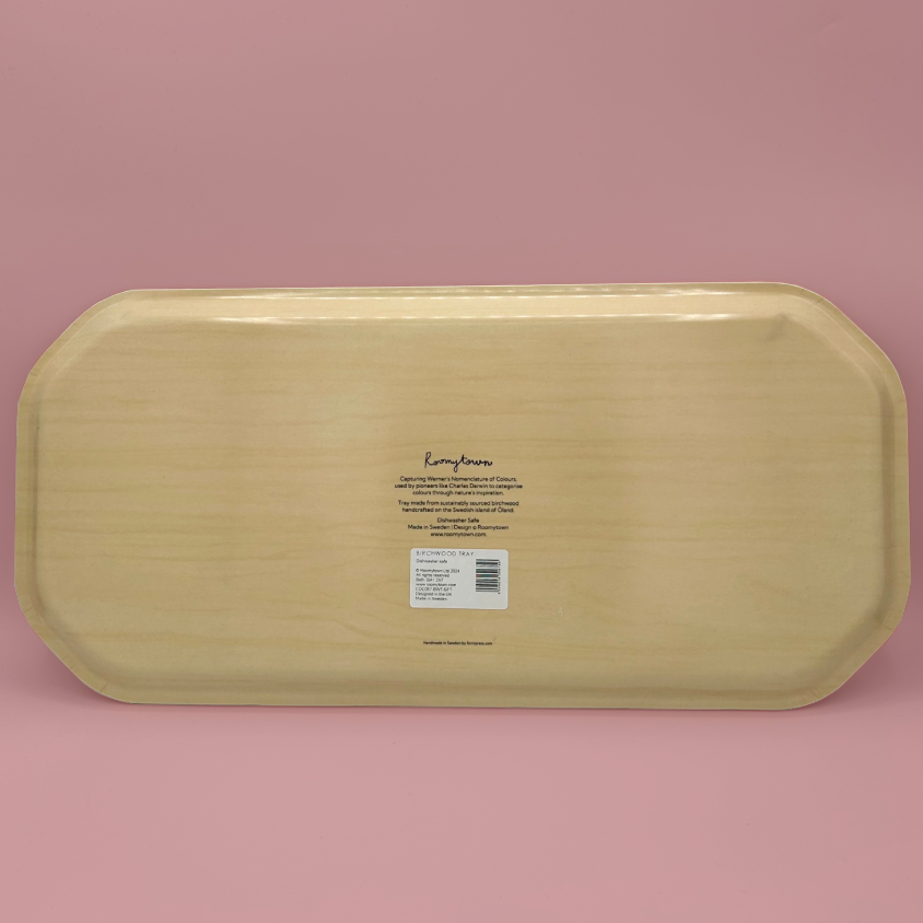 The underside of a light natural wood changing tray with rounded corners, featuring printed product information and a barcode sticker, set against a solid pink background.