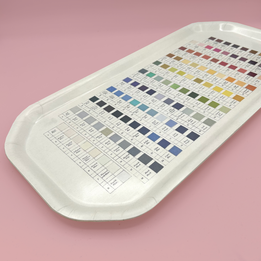 A wooden tray printed with a comprehensive color chart showing various paint or pigment swatches in rows, set against a solid pink background.