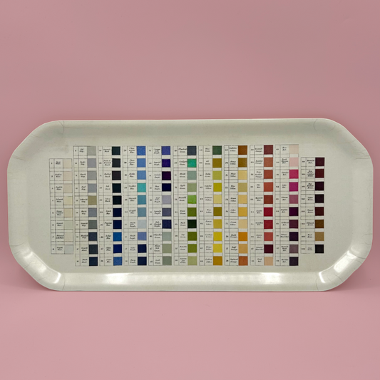 A rectangular colour chart tray with rounded corners, displaying multiple small color swatches arranged in columns with labels and numbers, set against a solid pink background.