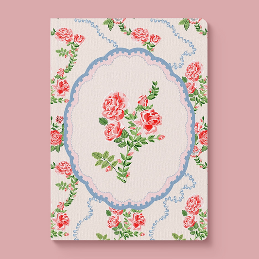 A hardcover notebook with a vintage floral design, featuring pink roses and green leaves around an ornate blue and pink oval frame on a cream background, set against a solid dusty pink backdrop.