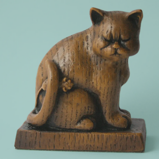 A carved cat figurine with a wooden finish, sitting on a rectangular wooden base against a solid light teal background.