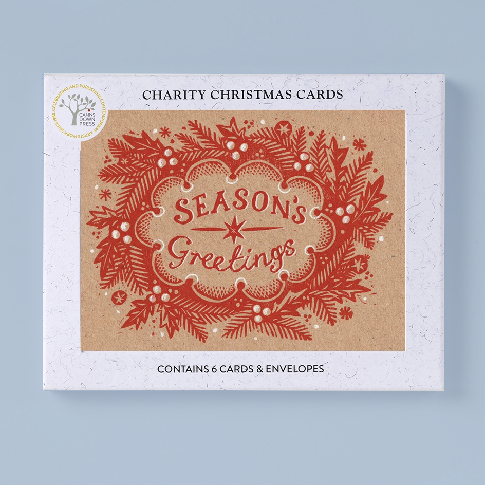 A rectangluar product box for charity Christmas cards, shown on a light blue background. The box front features a kraft-paper style central panel with a red ornate holiday wreath and the words 'Season's Greetings,' plus black text reading 'Charity Christmas Cards' and 'Contains 6 cards & envelopes.