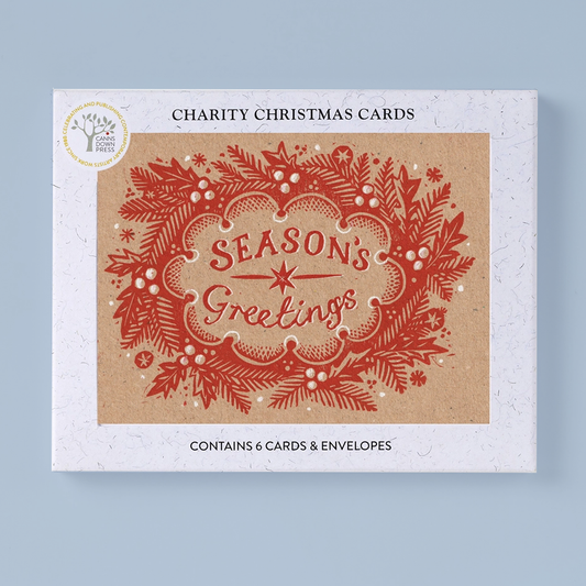 A rectangluar product box for charity Christmas cards, shown on a light blue background. The box front features a kraft-paper style central panel with a red ornate holiday wreath and the words 'Season's Greetings,' plus black text reading 'Charity Christmas Cards' and 'Contains 6 cards & envelopes.