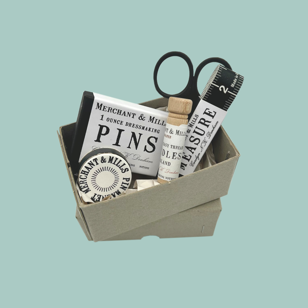 Box containing sewing supplies including pins, scissors, and measuring tape on a light blue background