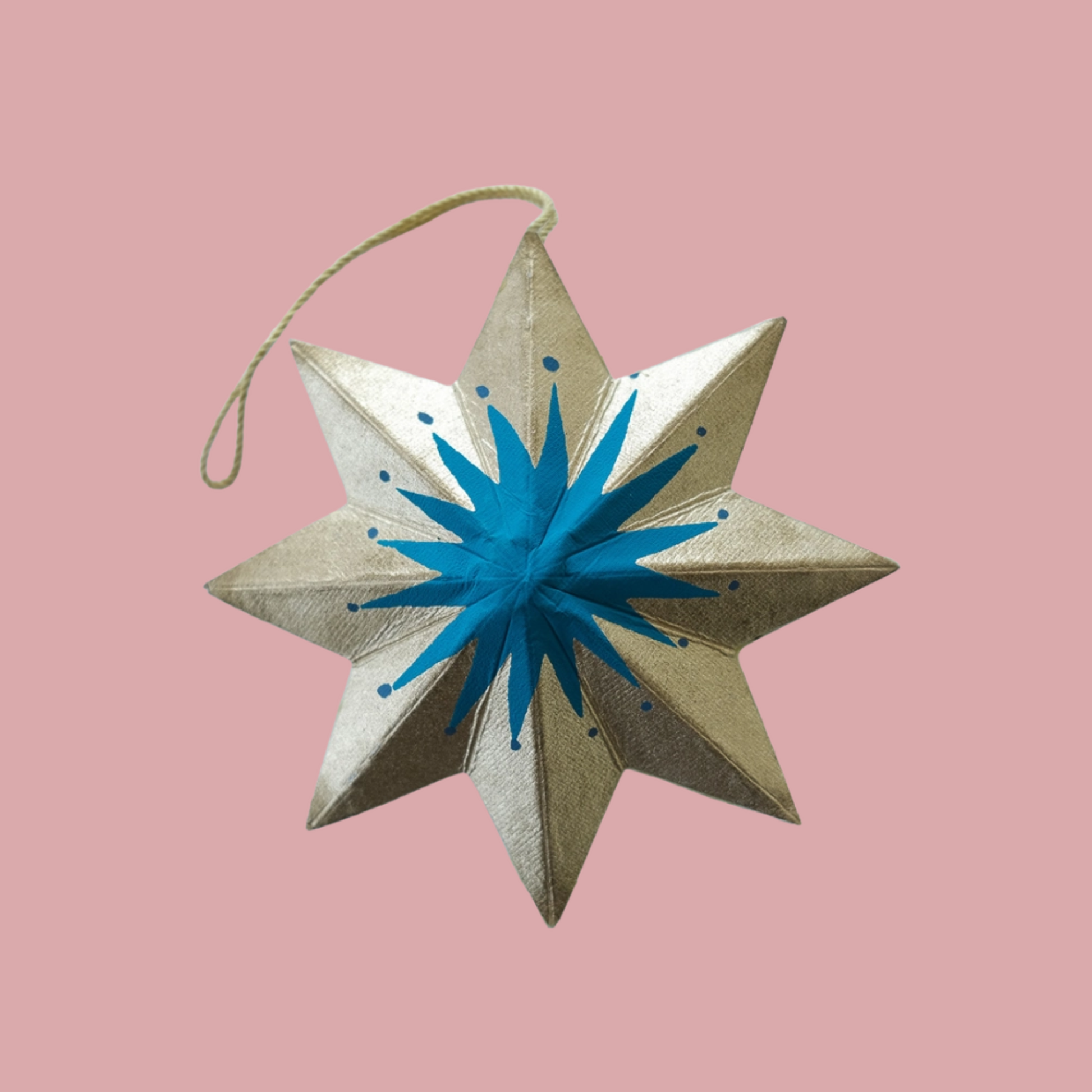 A metallic gold eight-pointed star ornament with a blue starburst center and a hanging string, set against a solid pink background.