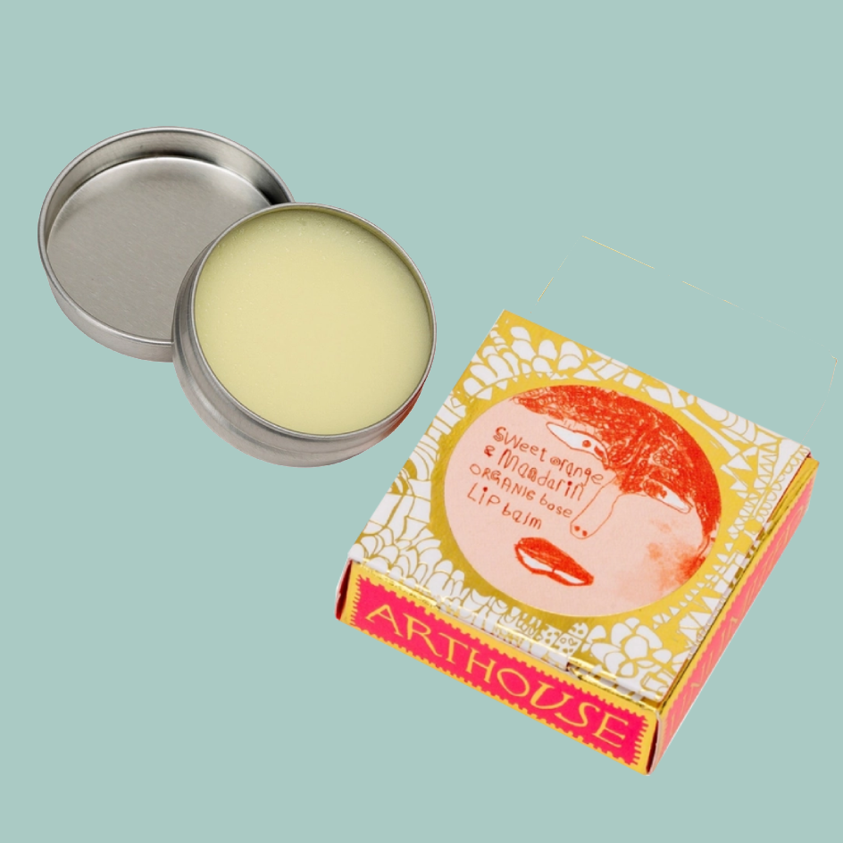 An open round metal tin of pale yellow balm next to its illustrated product box on a light blue background. The box shows a stylized face