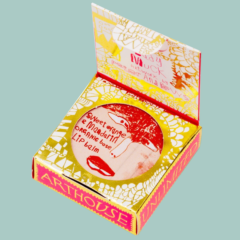 An open square box of Arthouse Unlimited lip balm, showing a round balm tin with red lips and handwritten text inside a gold and white patterned box on a pale blue background.