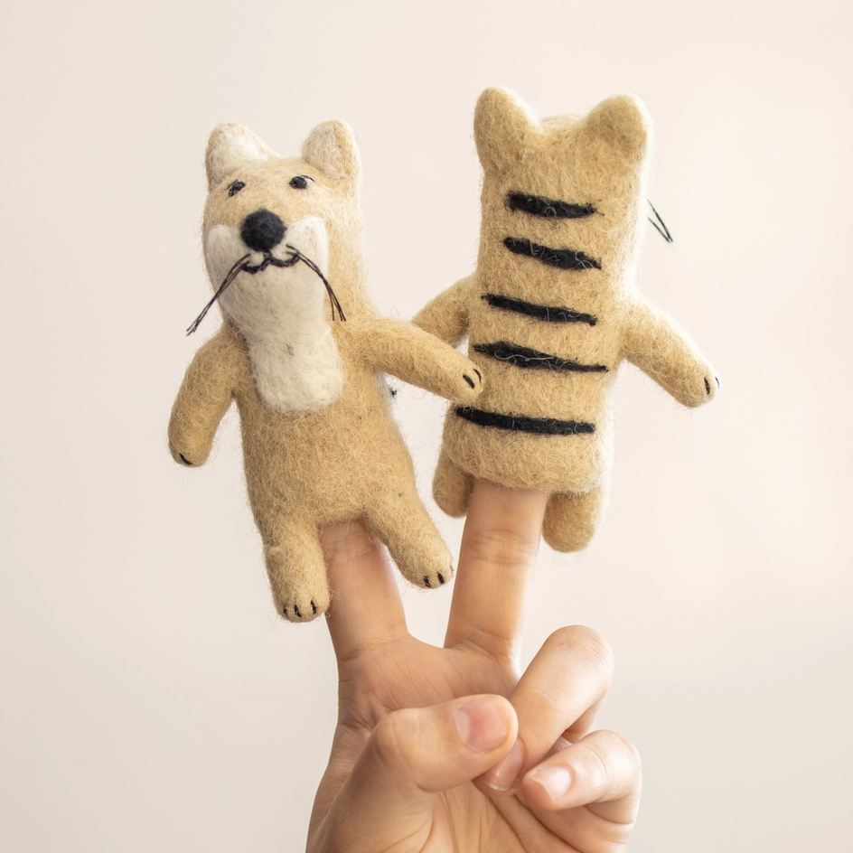 A hand holding two felt animal finger puppets, a tan tiger with a white chest and black nose on one finger, and a tan tiger with black stripes on the other finger, against a plain light beige background.