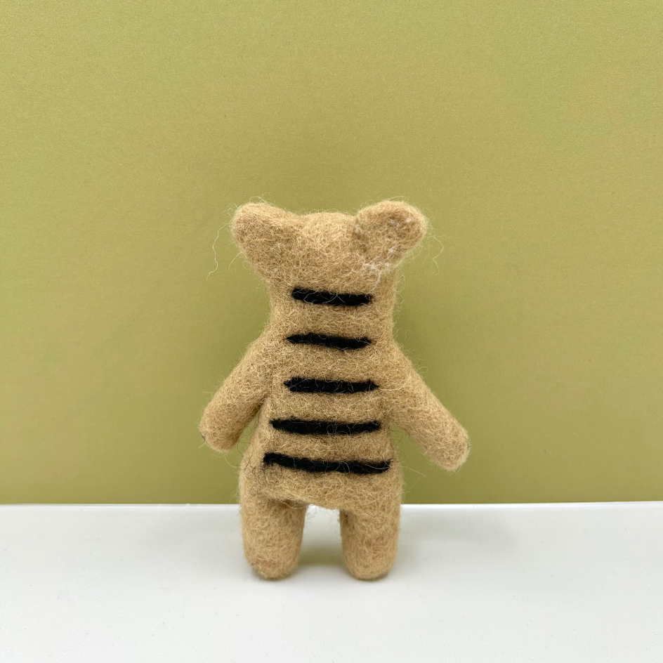 A small felt tiger toy shown from the back, made from beige wool with black stripes, standing on a white surface against a muted olive-green background.