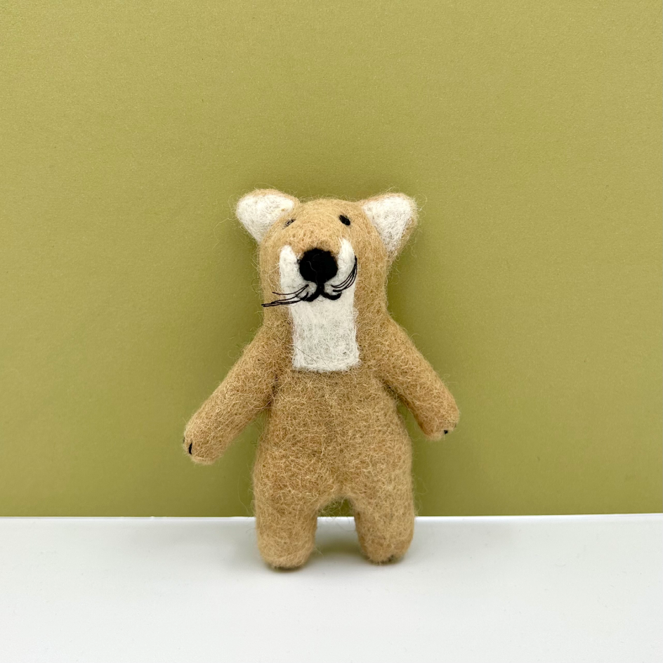 A small felted wool tiger with a tan body, cream inner ears and chest, and a black embroidered nose and whiskers, standing against a muted green background on a white surface.