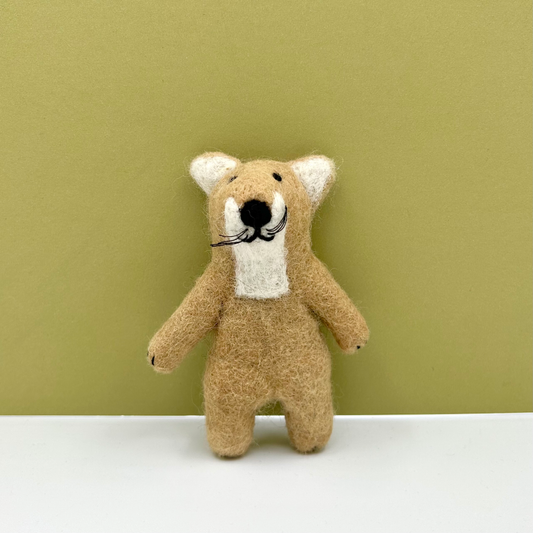 A small felted wool tiger with a tan body, cream inner ears and chest, and a black embroidered nose and whiskers, standing against a muted green background on a white surface.