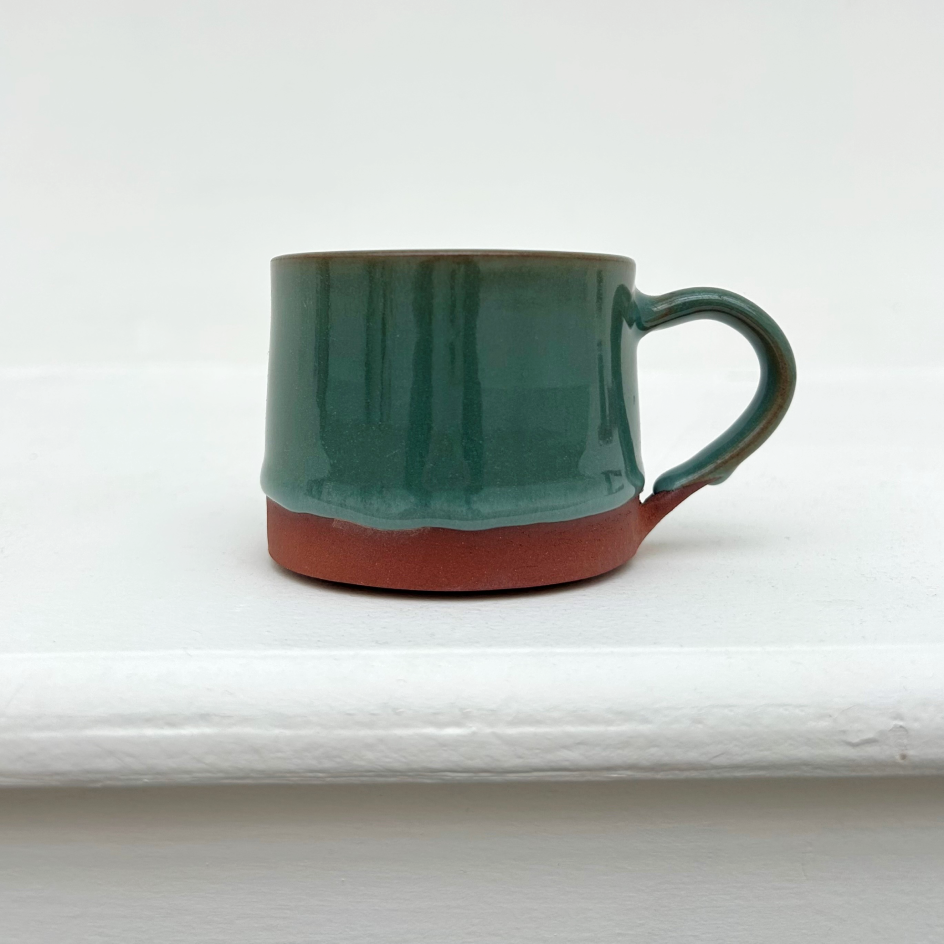 A ceramic mug with a terracotta coloured base and a teal glossy glaze. The mug is a on a white ledge with a white wall behind.