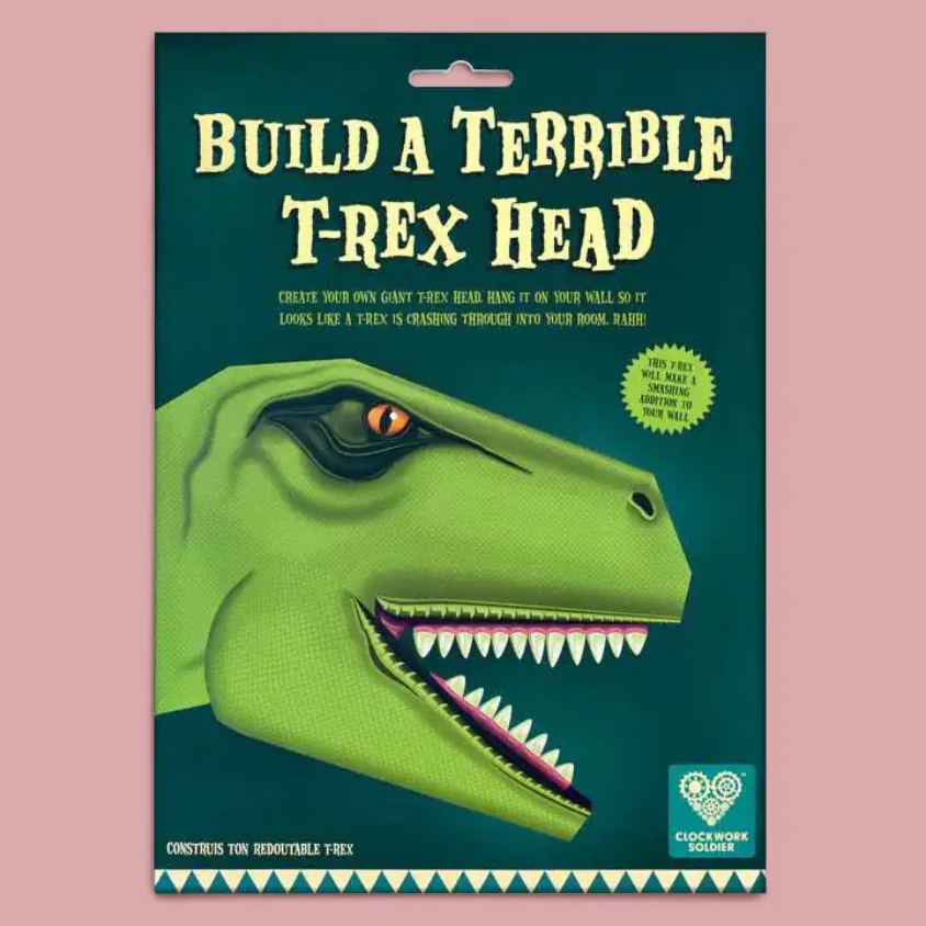 A  craft kit package titled 'BUILD A TERRIBLE T-REX HEAD' on a dusty pink background. The package features a green illustrated T-rex head.