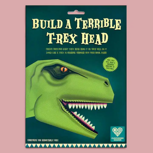 A  craft kit package titled 'BUILD A TERRIBLE T-REX HEAD' on a dusty pink background. The package features a green illustrated T-rex head.