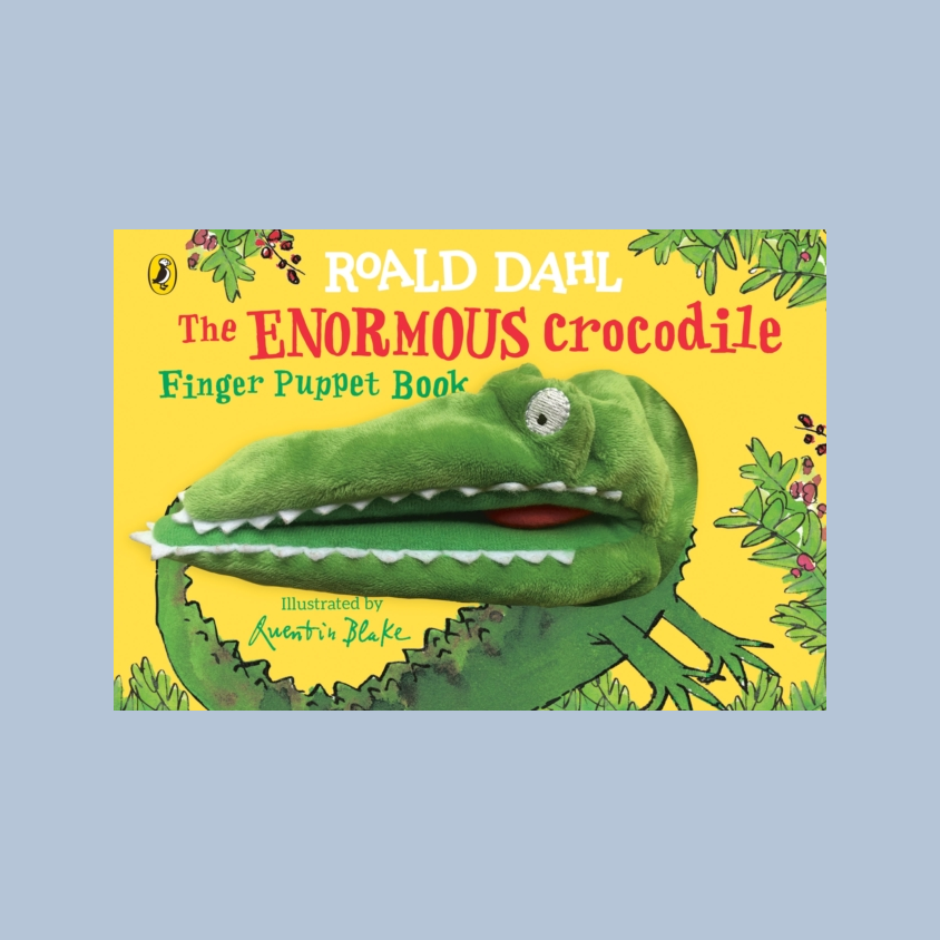 The front cover of the children's book 'The Enormous Crocodile' by Roald Dahl, featuring a large green puppet crocodile on a bright yellow background with illustrated foliage. The book is centered on a light blue-gray backdrop.