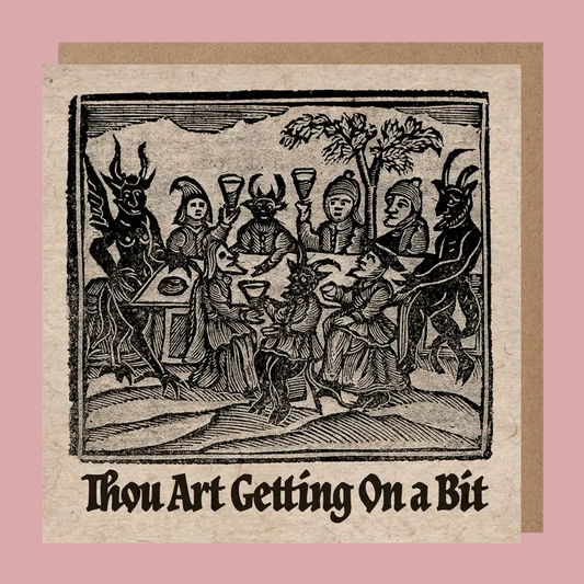 A square  greetings card with an illustration of medieval figures around a table, with the text 'Thou Art Getting On a Bit' below. The card is shown against a kraft paper enveloped on a pink background.