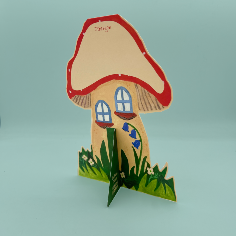 A mushroom-shaped card with a red-capped mushroom cap, small blue window details, green grass, and bluebell flowers, photographed against a pale blue background.