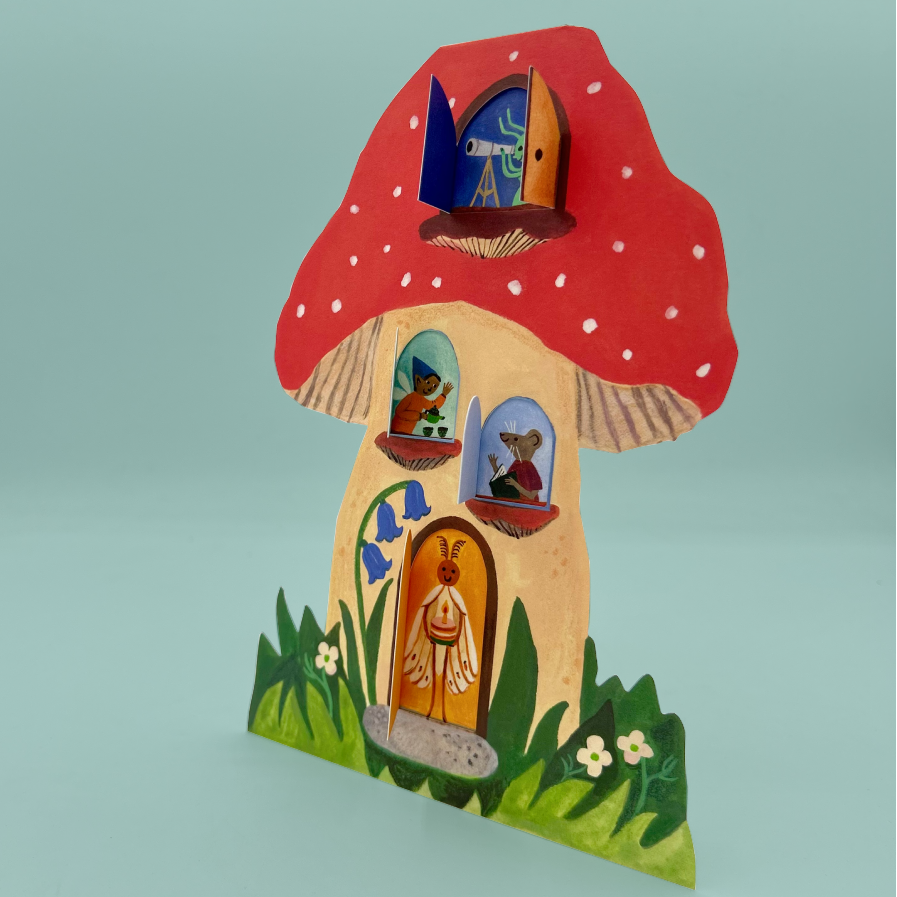 A greetings card shaped like a mushroom, featuring a red cap with white spots, a beige body, and colorful painted windows and door details, set against a solid light blue background.