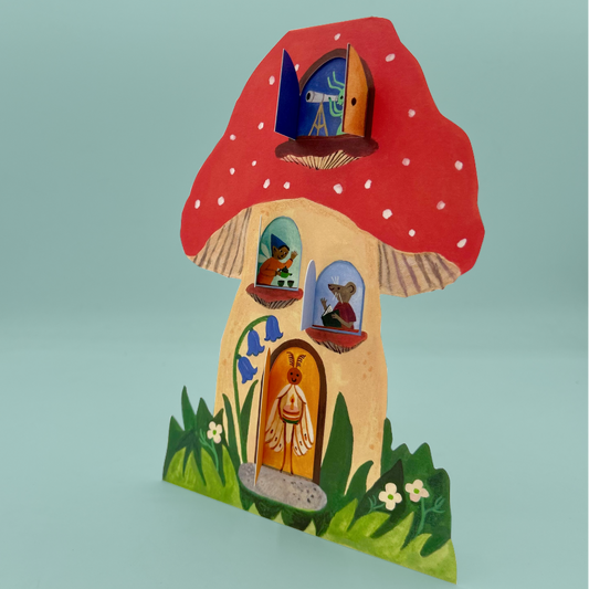 A greetings card shaped like a mushroom, featuring a red cap with white spots, a beige body, and colorful painted windows and door details, set against a solid light blue background.
