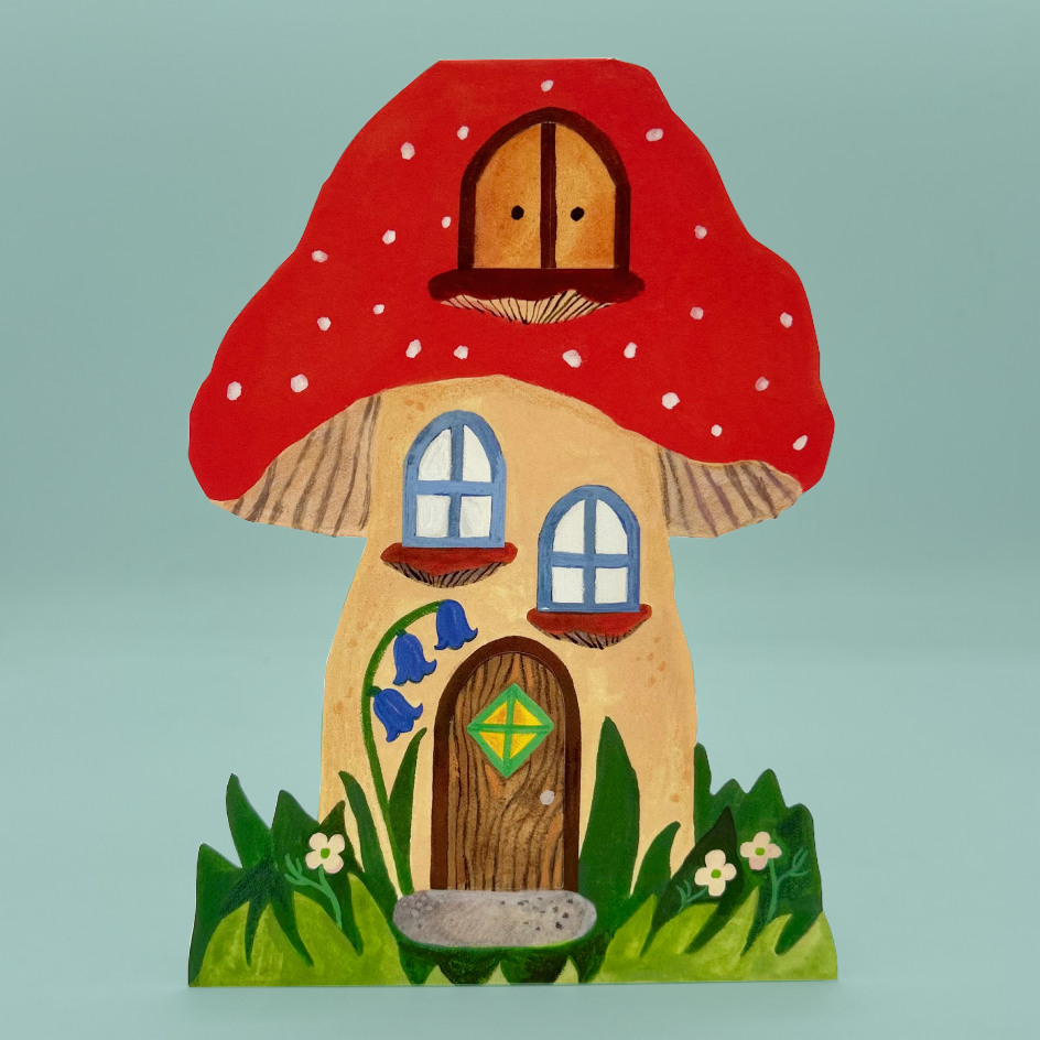 A greetings card shaped like a mushroom, featuring a large red cap with white polka dots, a beige body, a brown door with a green diamond, blue windows, and green grass at the base, set against a solid light blue background.