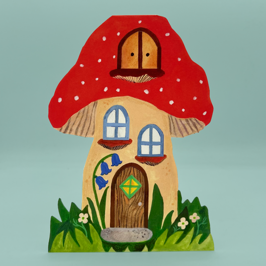 A greetings card shaped like a mushroom, featuring a large red cap with white polka dots, a beige body, a brown door with a green diamond, blue windows, and green grass at the base, set against a solid light blue background.
