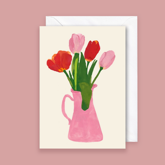 A greeting card with a watercolor-style illustration of tulips in a pink pitcher, shown with a white envelope against a dusty pink background.