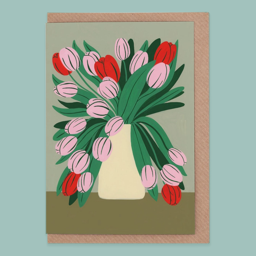 Greetings card of pink and red tulips in a cream vase, on a green background. The card is atop a kraft envelope on a blue background. 